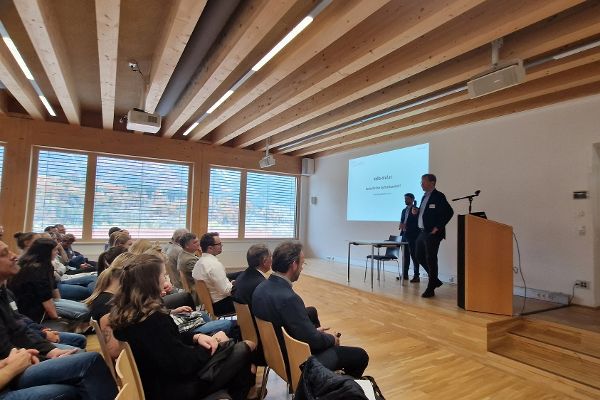 Die Eröffnung der Veranstaltung, Seitenansicht des Publikums mit Blick auf den Redner und die Präsentation, Fenster im Hintergrund mit Blick auf Wälder