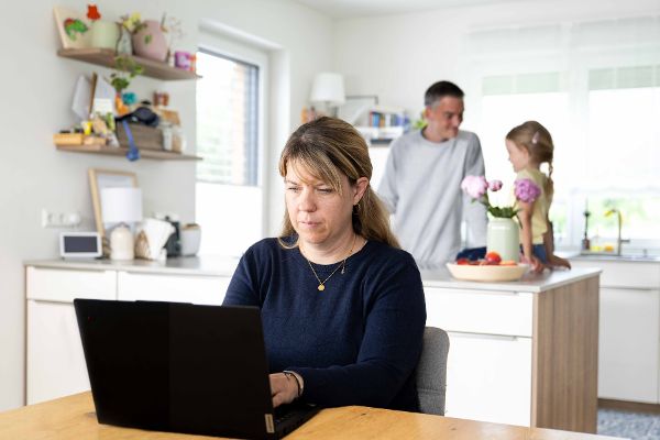 Eine Frau sitzt im Homeoffice am Laptop, im Hintergrund ist der Vater mit der Tochter in der Küche