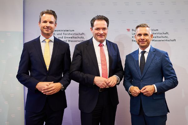 Drei Männer in Anzügen stehen nebeneinander vor einer Wand mit Text und Logos des Bundesministeriums für Landwirtschaft und Forstwirtschaft.