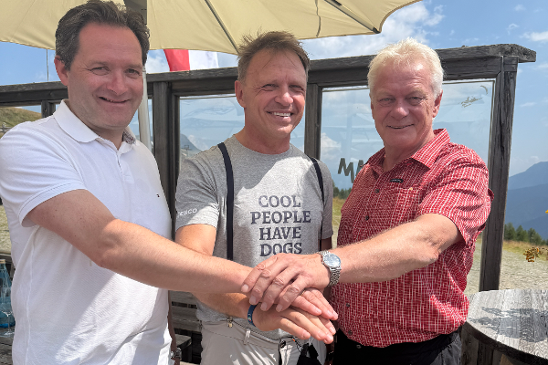 Drei Männer stehen unter einem Sonnenschirm und legen ihre Hände übereinander, einer trägt ein T-Shirt mit der Aufschrift 'COOL PEOPLE HAVE DOGS'