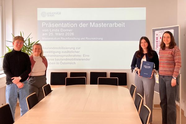 Vier Personen stehen in einem Besprechungsraum vor einer Präsentationsfolie, eine Person hält eine gebundene Arbeit in der Hand