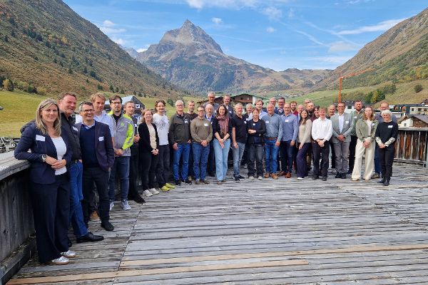 Gruppenfoto der Workshop Teilnehmerinnen und Teilnehmer auf einer Holzterrasse vor Berglandschaft mit spitzem Gipfel und Häusern im Tal