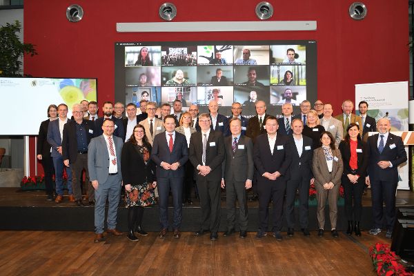 Gruppenfoto der Teilnehmer:innen an der woodPoP-Veranstaltung inklusive der Online-Teilnehmer im Hintergrund auf einer LED-Wand. Landwirtschaftsminister Totschnig steht zentral in der ersten Reihe.