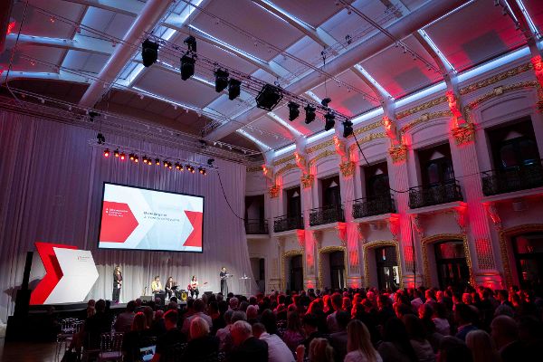 Blick über die Publikumsreihen nach vorne zu einer Bühne in einem prunkvollen Saal. Auf der Bühne sitzen drei Musiker:innen, links steht eine Moderatorin und rechts steht ein Jurymitglied und spricht ins Mikro. Auf einer Projektion im Hintergrund sieht man das Logo des Staatspreis Holzbau.