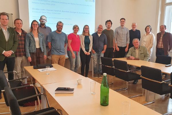 nebeneinander gereihte Menschen in einem in U-Form angneordneten Besprechungstischen, im Hintergrund eine Praesentationsabbildung dieser Arbeitsgruppe