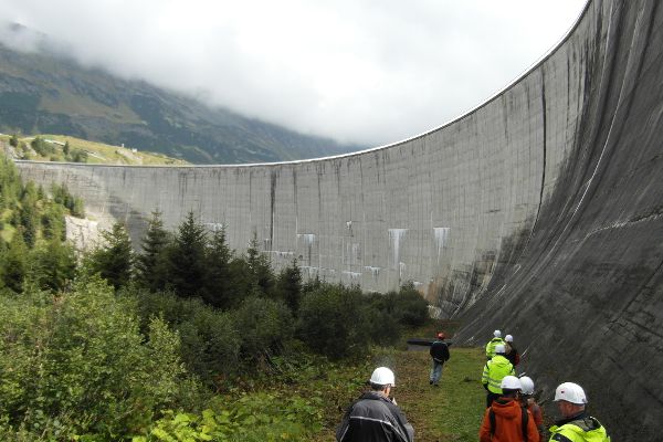 Staumauer Sperre Kops - die Kursteilnehmer gehen am Fuß der Gewölbesperre auf einem schmalen Streifen, daneben Strauch- und Baumbewuchs
