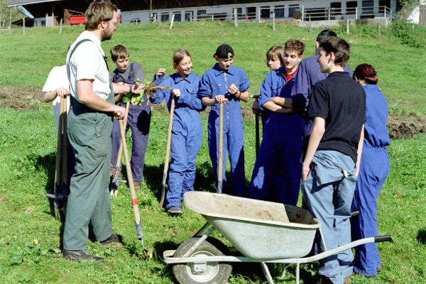 Lehrer, Schülerinnen und Schüler einer Fachschule stehen auf einer Wiese, Scheibtruhe im Vordergrund