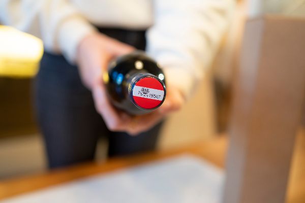 Person hält eine Weinflasche mit rotem Verschluss und einem weißen Etikett mit dem Code 'DE 01 195417'