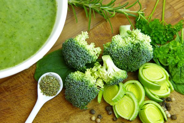 grüne Cremesuppe in einer Schüssel, daneben Gemüse, Kräuter und Gewürze