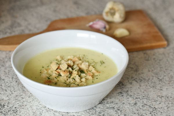 Schüssel mit heller, cremiger Suppe und Croutons, im Hintergrund Schneidebrett mit Knoblauch