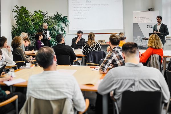 Teilnehmerinnen eines Expertinnenworkshops sitzen in einem Raum, Vortragender steht vor einer Präsentation an der Wand