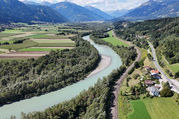 Luftaufnahme der Drau vor ihrer Revitalisierung in einem alpinen Tal. Der Fluss verläuft in einem breiten, vergleichsweise gleichförmigen Bett durch eine von Landwirtschaft geprägte Landschaft mit Feldern, Wiesen und bewaldeten Uferstreifen. Entlang des rechten Ufers verlaufen eine Bahnstrecke und eine Straße mit einzelnen Wohnhäusern. Bewaldete Berghänge und hohe Gipfel prägen die Kulisse unter einem teilweise bewölkten Himmel.