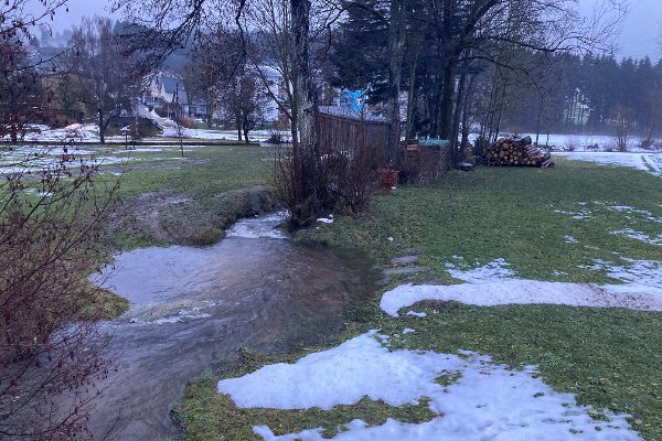 Fluss umrandet von gruener Wiese leicht Schnee bedeckt.