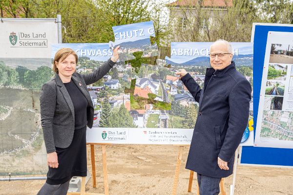 zwei Personen halten ein Puzelteil mit Aufschrift Schutz empor. Dahinter steht die Bautafel mit der Landschaftsaufnahme um den Gabriachbach