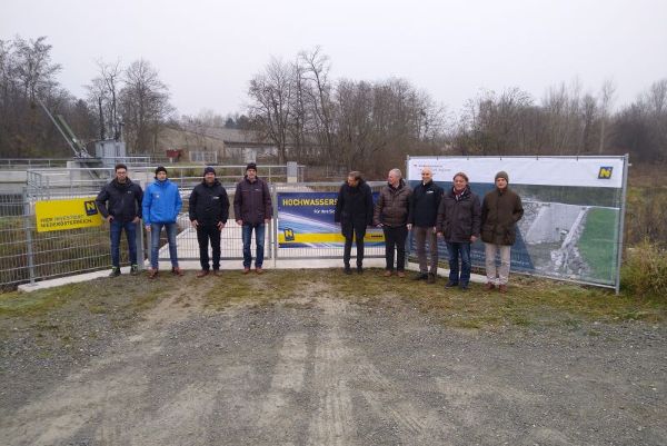 sieben Personen vor den Bautafeln und Bauzaun, im Hintergrund Baugelaende und Baeume und Haeuser