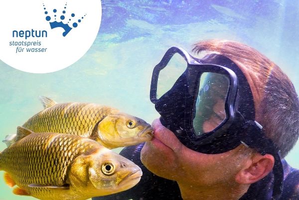 Das Hauptsujet des Neptun Staatspreis 2027 zeigt einen Mann mit Taucherbrille unter Wasser, umgeben von Fischen. Ein Fisch berührt ihn dabei sanft mit einem kleinen Kuss.