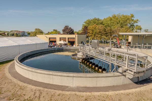 Die Kläranlage Podersdorf bei ihrer Eröffnung: Ein großes rundes Nachklärbecken mit Wasser und mechanischer Räumeranlage steht im Vordergrund. Im Hintergrund sind Betriebsgebäude, Festzelte und Besucherinnen und Besucher zu sehen.