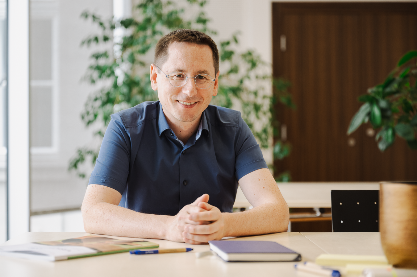 Mann sitzt mir verschränkten Armen am Tisch und lächelt in die Kamera