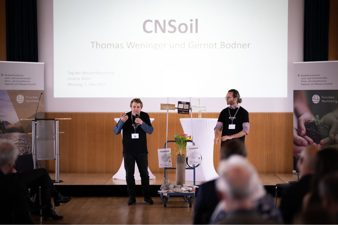 Gernot Bodner und Thomas Weninger stellen ihr Projekt dem Publikum des "Tag der Ressortforschung 2025" vor. Die beiden stehen vor der Leinwand, haben einen Modellaufbau ihres Versuchs zwischen sich, das Publikum sieht man nur von hinten
