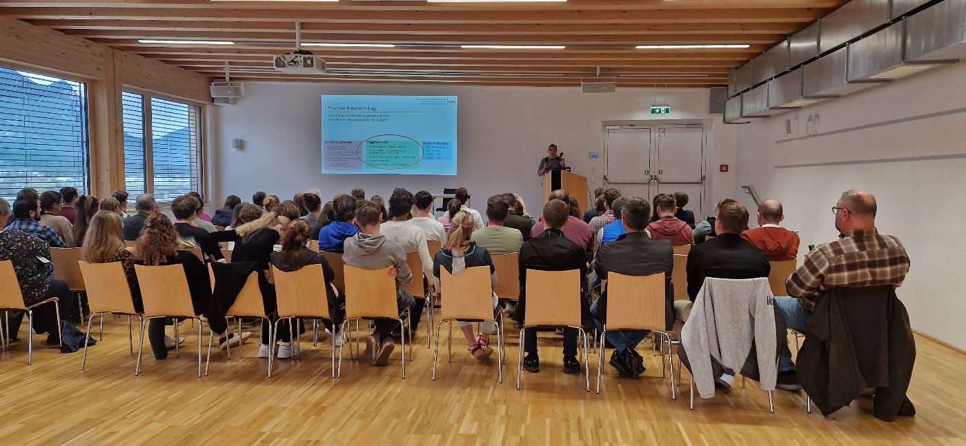 Blick von hinten auf das sitzende Publikum, welches einem Vortrag auf der Bühne lauscht