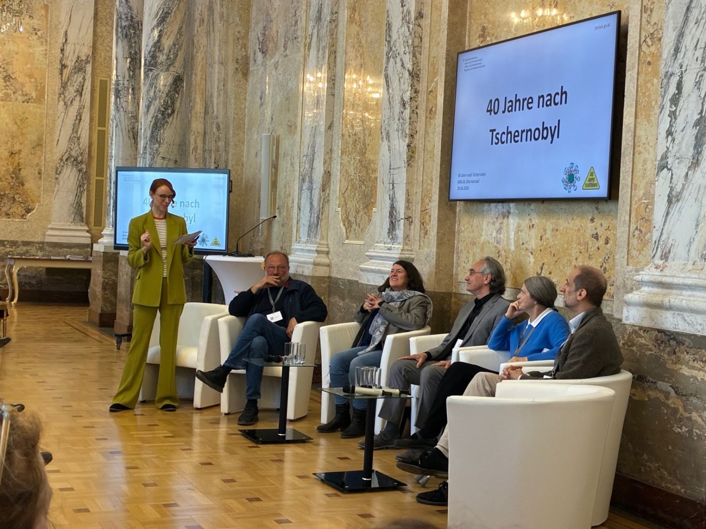 Am Foto sieht man die Moderatorin stehend und die Teilnehmer:innen an der Podiumsdiskussion im Sessel sitzend