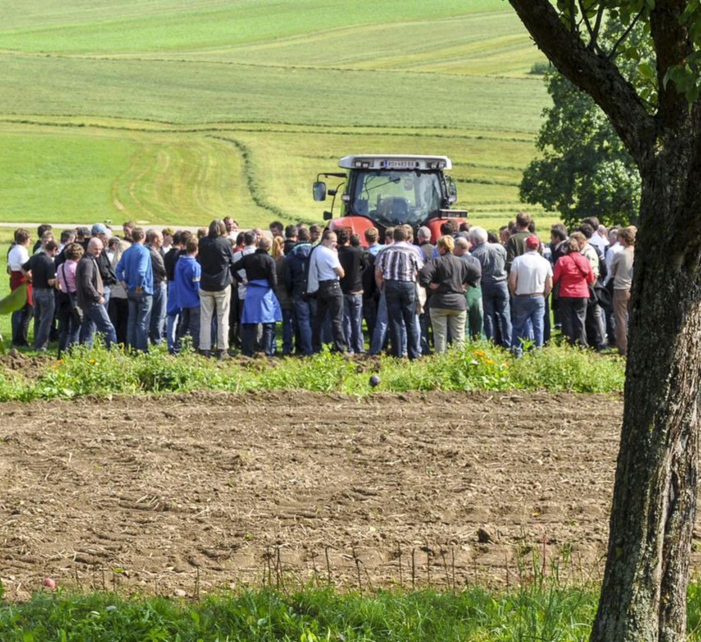 Gruppe von Personen steht in Agrarlandschaft vor einem Traktor. Im Vordergrund eine Ackerfläche, im Hintergrund Grünland.