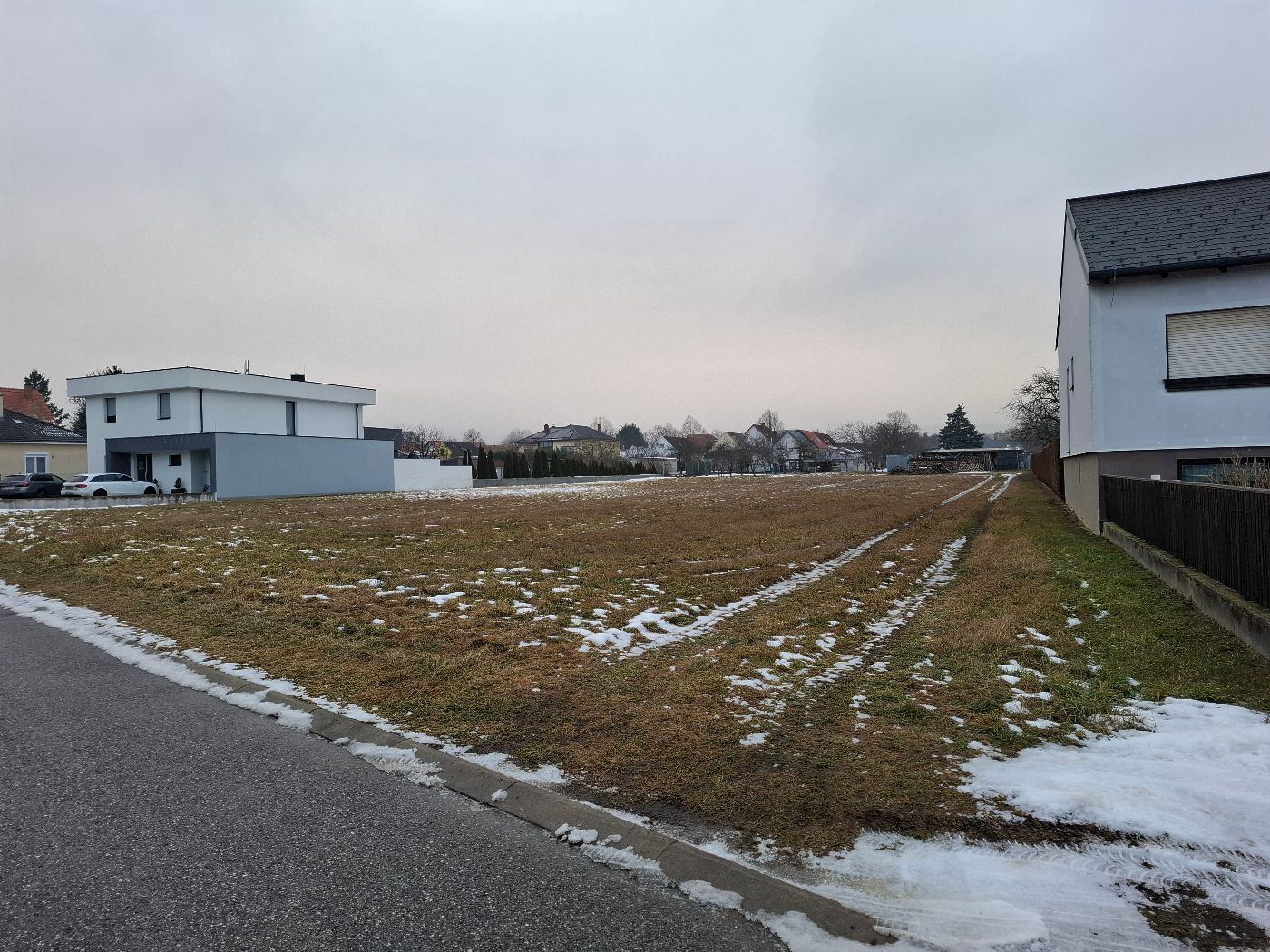 Freies Bauland mit leicht schneebedecktem Gras zwischen zwei Wohnhäusern unter bewölktem Himmel