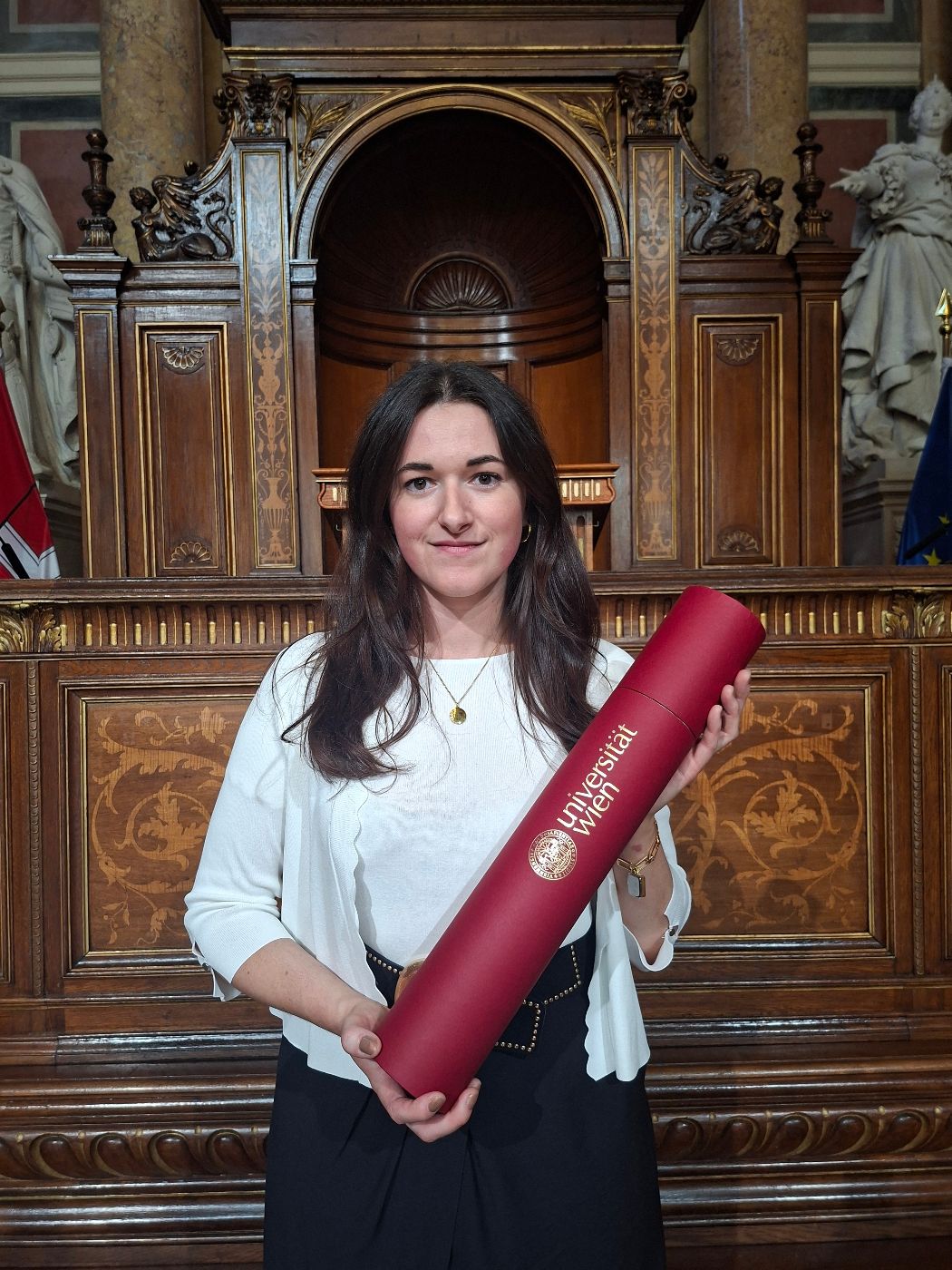 Frau steht vor einer verzierten Holzvertäfelung in einem historischen Saal und hält eine rote Urkundenrolle mit dem Schriftzug 'Universität Wien'