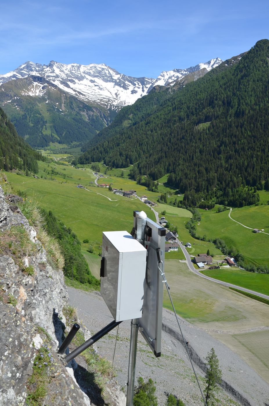 Ein silbernes, rechteckiges Messgerät steht an einem Berghang. Dahinter sieht man eine Straße und eine bergige Landschaft.