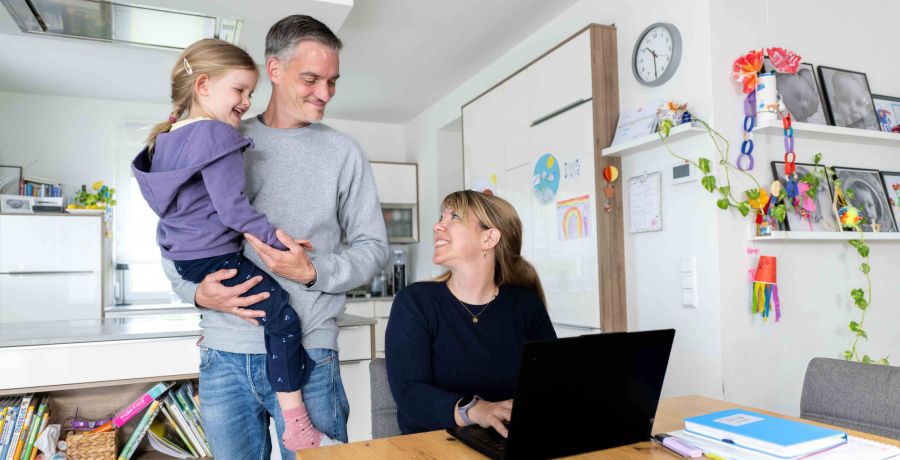 Eine Frau sitzt im Homeoffice vor ihrem Laptop, daneben trägt der Mann die Tochter auf dem Arm.