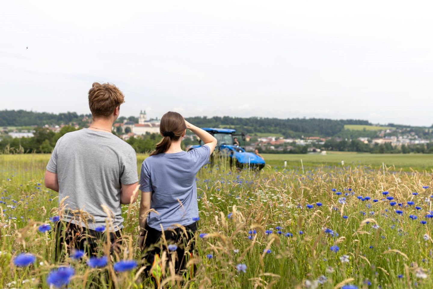 Eine Schülerin und ein Schüler stehen mit dem Rücken zum Fotografen auf einem Feld und schauen zu einem Traktor