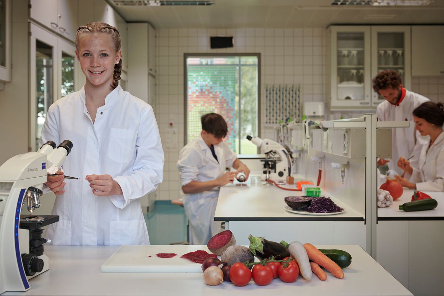 Im Vordergrund ein MÄdchen mit Laborkittel in einem Labor, das vor einem Mikoskip steht. Im Hintergrund sind weitere Schüler zu sehen.