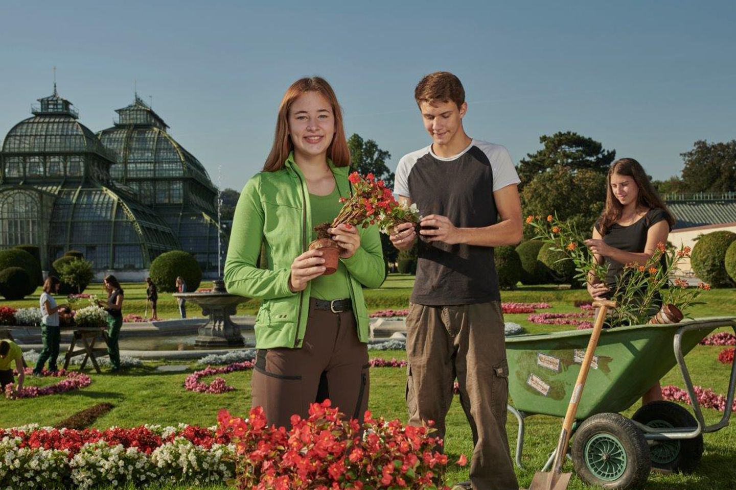 Zwei Mädchen und ein Junge mit Blumen und Gartenarbeitsgeräten. Im Hintergrund sind die Palmenhäuser zu sehen.