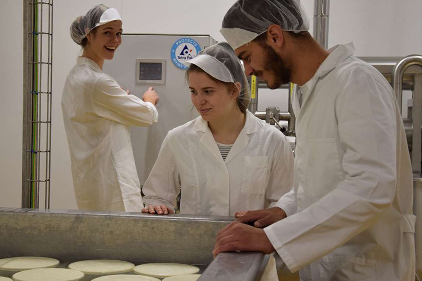 Ein Bursch und zwei Mädchen in weißer Schutzkleidung bei der Herstellung von Käse