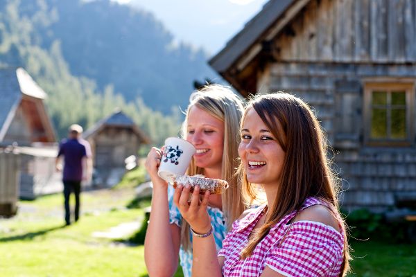 Zwei Wanderinnen genießen die kulinarischen Köstlichkeiten auf einer Almhütte im Freien.