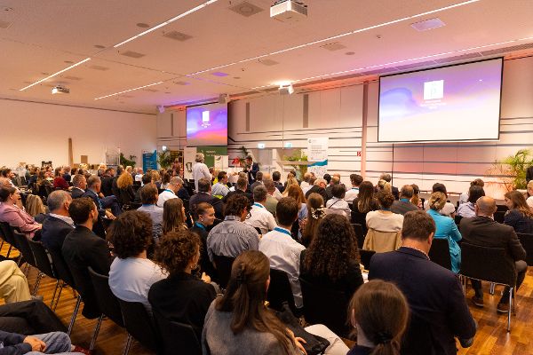 Großer Saal mit Publikum, das auf eine Präsentation auf zwei Leinwänden schaut