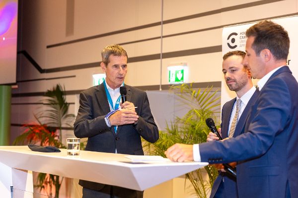 Drei Männer in Anzügen stehen an einem weißen Stehtisch, zwei halten Mikrofone, einer spricht, im Hintergrund Pflanzen und ein Notausgangsschild.