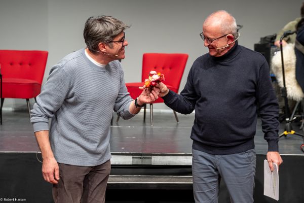 Zwei männliche Personen halten einen Trockenblumenstrauß und witzeln herum. Im Hintergrund knallrote Stühle.