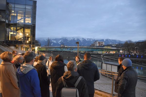 Eine Gruppe Menschen die an einer Stadtführung in Villach teilnehmen, im Hintergrund ein Bergpanorama, eine Brücke und ein Fluss