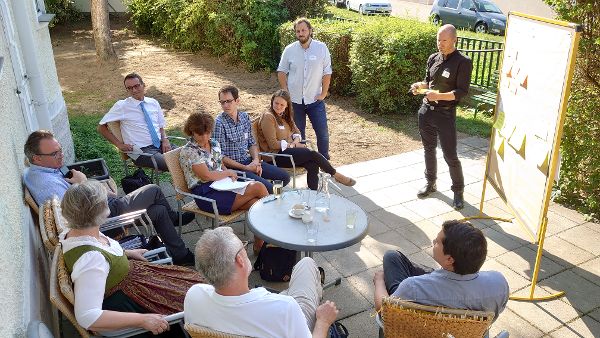 Teilnehmerinnen und Teilnehmer sitzen im Halbkreis und diskutieren. Der Workshopleiter steht am Pinboard und notiert das gesagte mit.