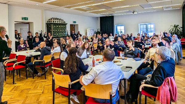 An der Ortskernwerkstatt teilnehmende Menschen sitzen an Tischen in einem großen Raum und diskutieren. 