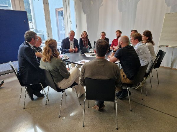 Mehrere Personen sitzen an einem Tisch in einem hellen Raum und diskutieren miteinander. 