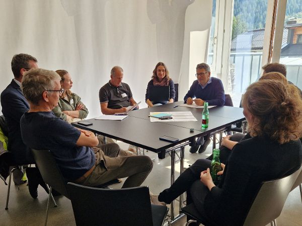 Gruppe von acht Personen sitzt an zwei zusammengestellten Tischen in einem hellen Raum mit großen Fenstern und unterhält sich