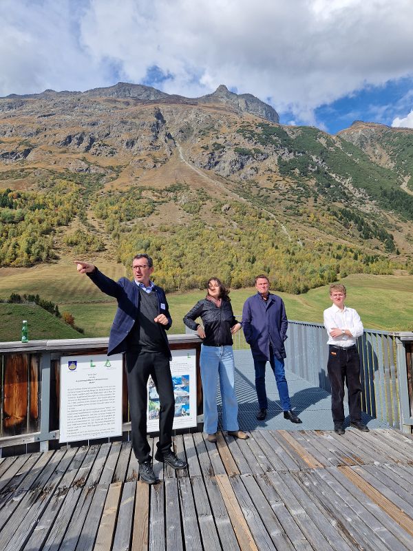 Vier Personen stehen auf einer Holzterrasse vor einem Bergpanorama, eine Person zeigt mit ausgestrecktem Arm nach links
