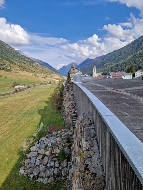 Blick von einer Mauer auf ein Tal mit Wiesen, Bergen und einem Dorf mit Kirchturm unter blauem Himmel mit Wolken
