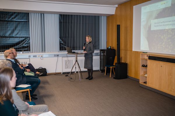 Sektionschefin Elfriede Moser steht in einem Raum an einem Stehtisch und spricht in ein Mikrofon. Vor ihr sitzen einige Menschen im Publikum. Hinter ihr sieht man eine an die Wand projizierte Präsentation.