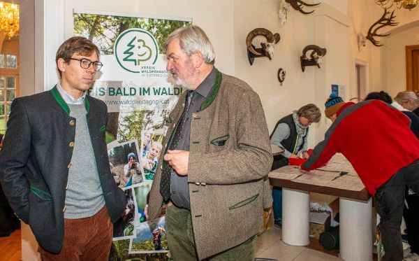 zwei Personen im Gespräch, im Hintergrund weitere Personen, die sich bei einem Tisch über Fachinformationen austauschen