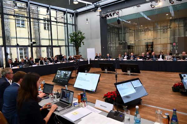 Gesamtansicht des Raumes. Die Teilnehmerinnen und Teilnehmer sind an Tischen in Form eines U platziert, dahinter sitzen weitere Personen. Auf der freien Fläche in der Mitte befinden sich zahlreiche Bildschirme, auf denen die Online-Teilnehmer zu sehen sind. 