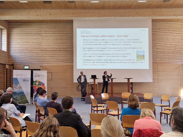Petra Völkl hält einen Vortrag auf der Bühne des Regional Policy Lab. Hinter ihr an der Wand die Projektion der Powerpoint-Präsentation, im Vordergrund sitzen einige Personen als Publikum.