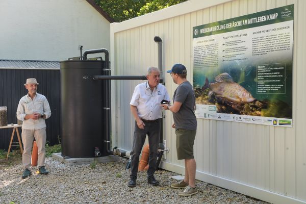 zwei Maenner unterhalten sich, seitlich ein Plakat mit Schrift und einem Fisch, im Hintergrund ein weitere Person